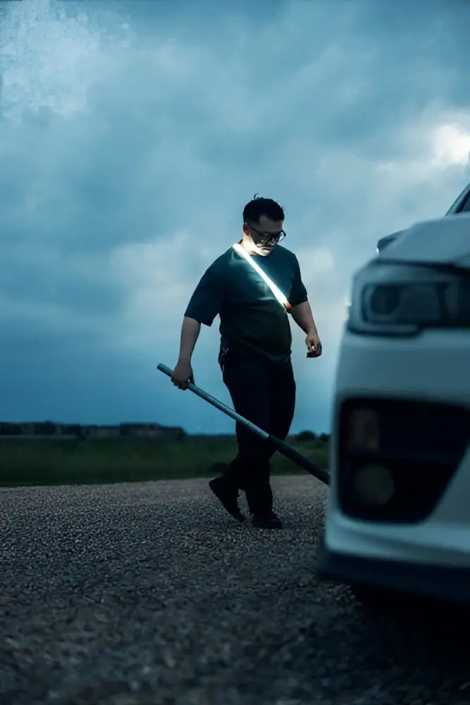 Mies tummissa vaatteissa ja silmälaseissa kävelee valkoisen auton sivulla kantaen bando 1k strap light työvaloa pitkällä kahvalla ja valolla