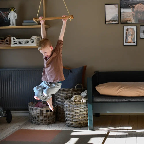 A child hangs from a bone oak green wooden monkey bar with rope suspension in a cozy neutrally toned room