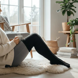 Pitkät villasukat beige neulepuseron ja mustien sukkahousujen kanssa istuvalla henkilöllä valkoisella matolla