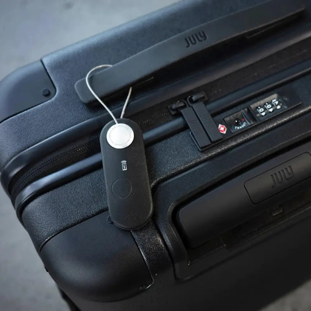 Musta suorakaiteen muotoinen scout travel luggage tag hopeisella pyöreällä napilla ja july-brändillä